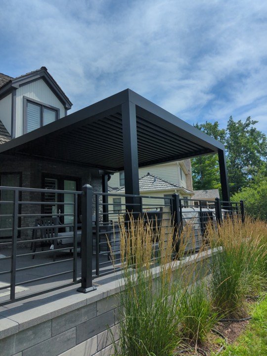 Modern louvered pergola with dark framing beside a cable-rail deck and planting beds.